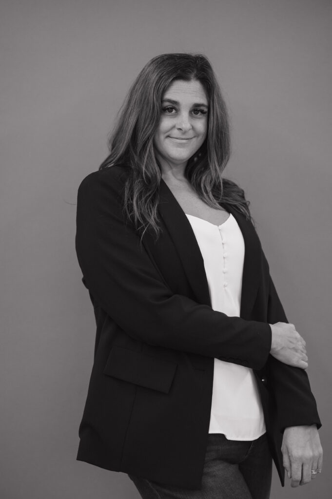 Black-and-white portrait of Jennifer Hume in a black blazer over a white top, one hand resting on her forearm, smiling softly