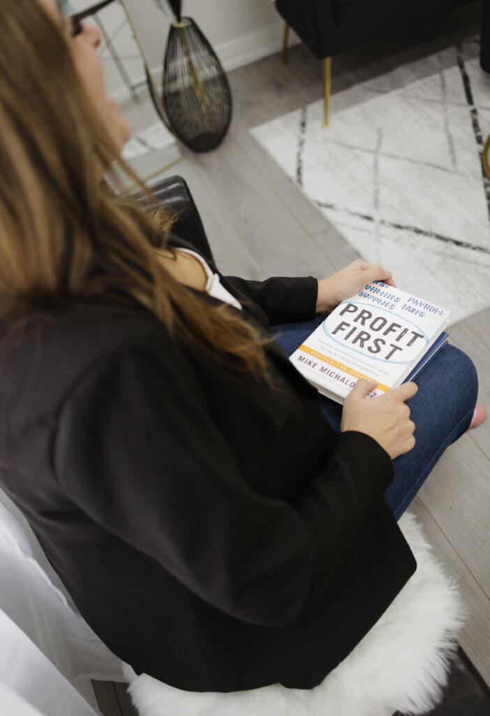 Jennifer Hume seated in a black blazer, seen over her shoulder, holding a copy of the book Profit First on her lap