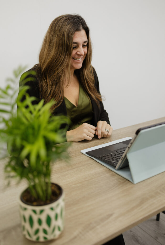 Jennifer Hume smiling at a tablet with a keyboard on a wooden desk, with a green potted plant in the foreground