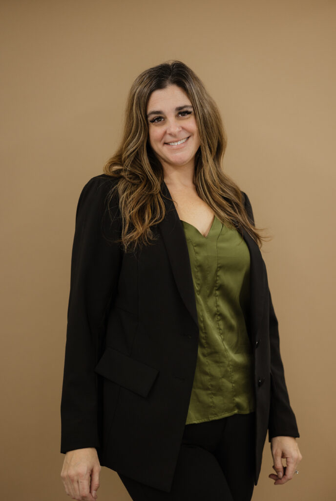 Jennifer Hume smiling, standing in a black blazer over an olive satin top against a warm tan background